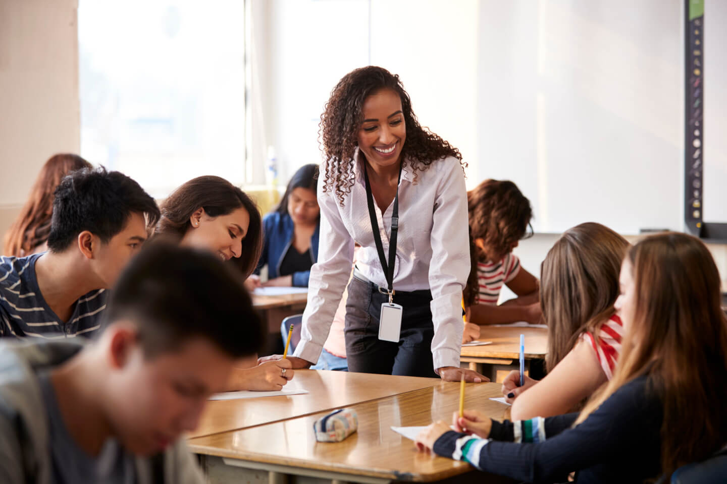 Female High School Teacher Standing By Student Table Teaching Lesson