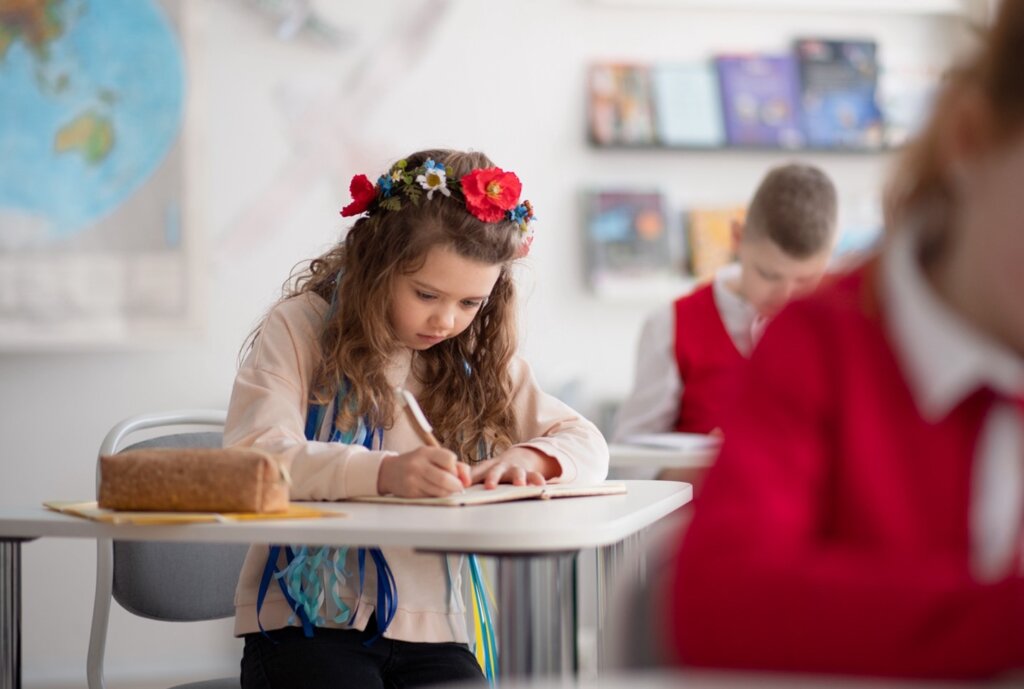 A young girl with a flower crown writing in her notebook in a classroom, surrounded by focused classmates.