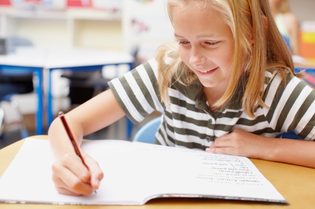 A young girl smiling while writing in her notebook in a cheerful classroom setting.