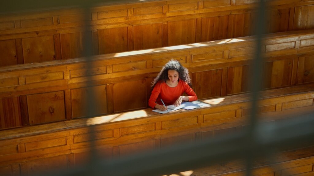 A student writes in a notebook in a bright, empty lecture hall with wooden benches.