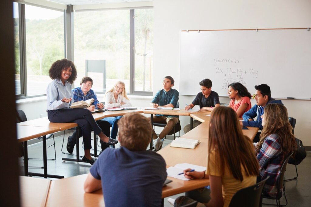 A teacher leading a discussion with high school students seated in a semi-circle classroom setting.