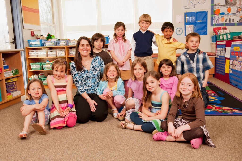 A smiling teacher posing with a group of young students in a colorful kindergarten classroom.