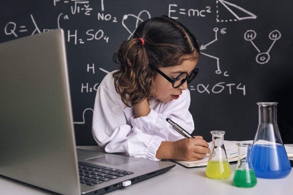 Young girl in lab coat writing in notebook with science beakers and formulas on chalkboard.