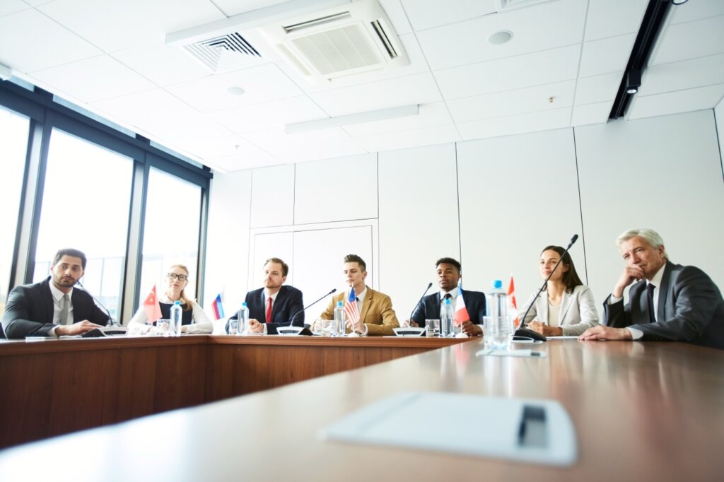 International panel of business professionals at a formal conference table with country flags and microphones.