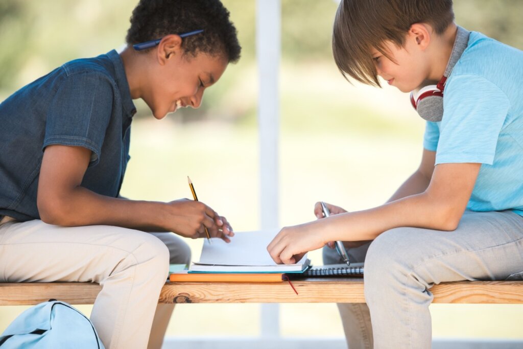 Two middle school boys sitting on a bench outdoors, collaborating on homework with notebooks and pencils.