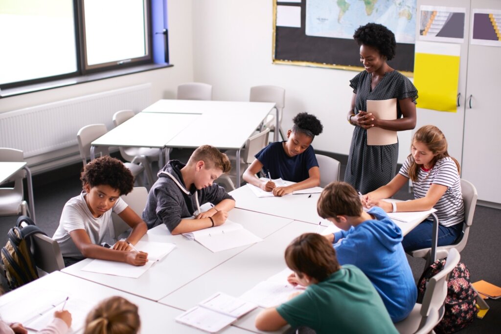 Diverse group of students writing at desks in a classroom while their teacher observes