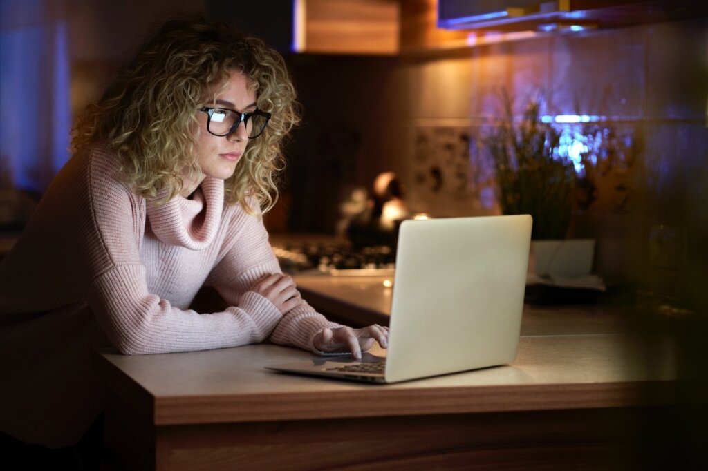 Young woman with curly blonde hair wearing glasses works late on a laptop in a warmly lit kitchen.