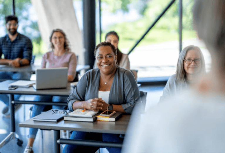 Adults seated at tables in a classroom, engaged in professional learning with books, laptops, and notebooks.