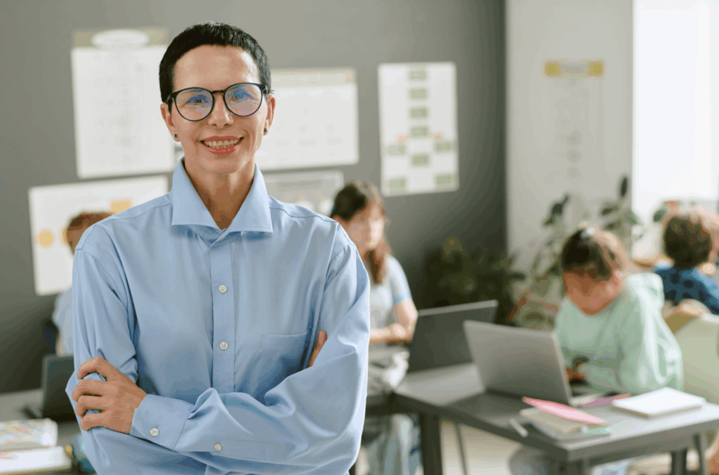 Confident teacher smiling in a modern classroom while students work on laptops in the background.