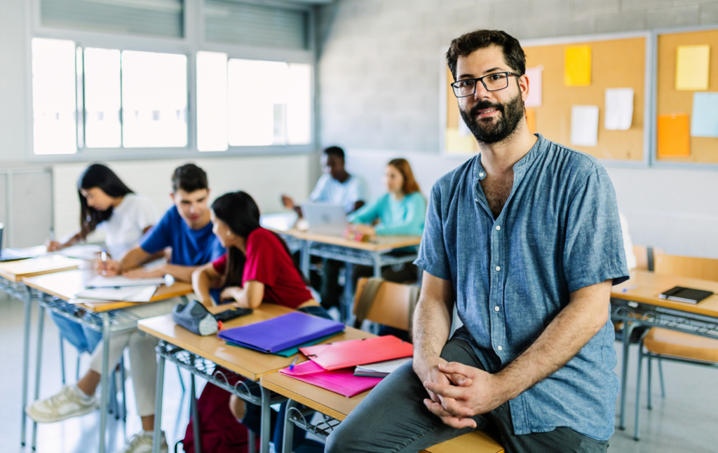 Male teacher sitting at the front of a classroom while students collaborate and work on assignments at desks behind him.